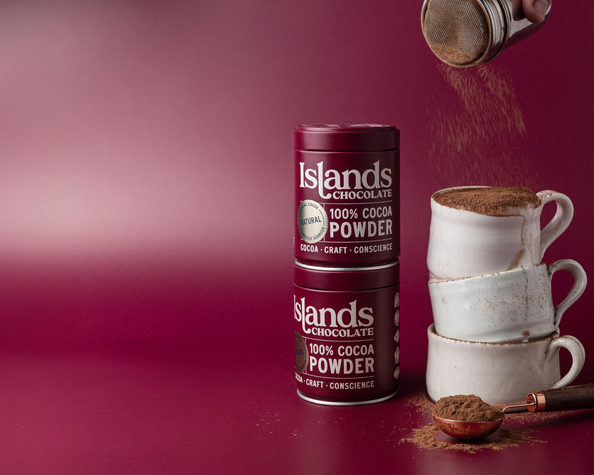 Two tins of Islands Chocolate 100% Cocoa Powder on a deep burgundy background, with stacked white ceramic mugs filled with cocoa, a spoon of cocoa powder, and cocoa being poured from a metal container.